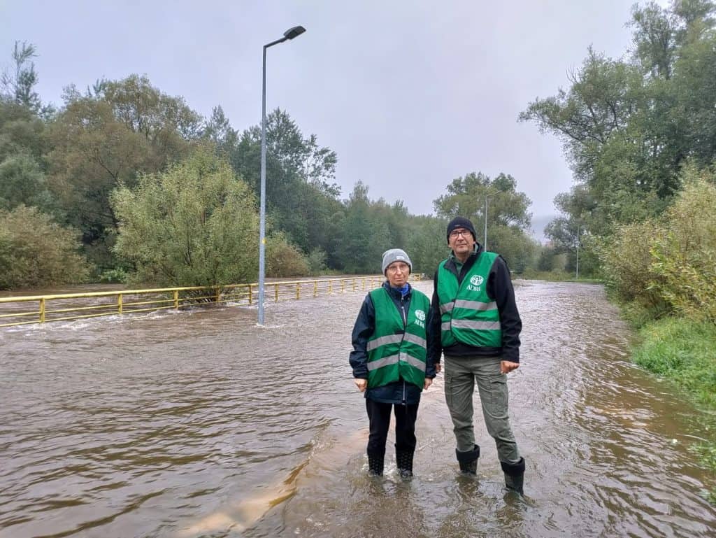 Two ADRA workers, a man and a woman, in teal ADRA vests stand in flooded waters