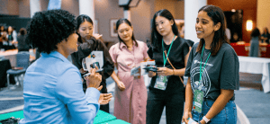 A young woman with curly hair and a blue shirt is standing a booth, talking to 4 other young women about ADRA