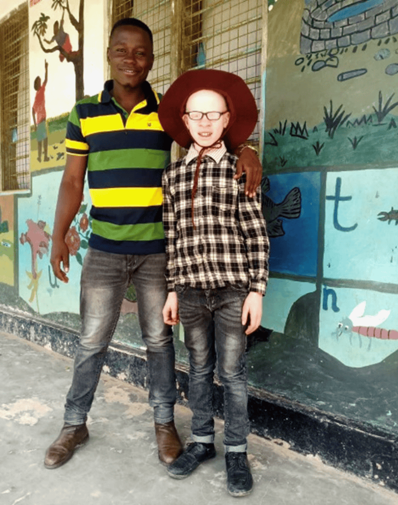 A man from Tanzania, in a striped yellow and green shirt, stands with a young boy with albinism wearing a cowboy hat and plaid shirt, outside the school