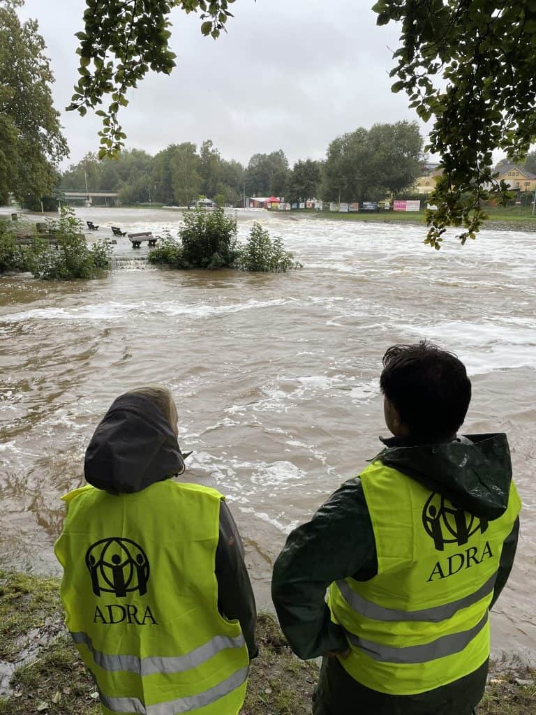 Two ADRA workers in bright neon yellow vests stand looking out over a flooding river.