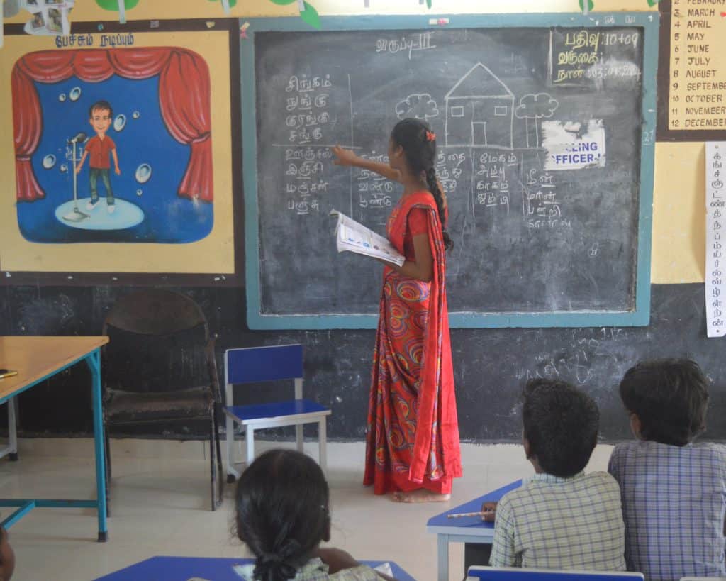 In India, a young woman in a pink and  yellow sari writes on a blackboard