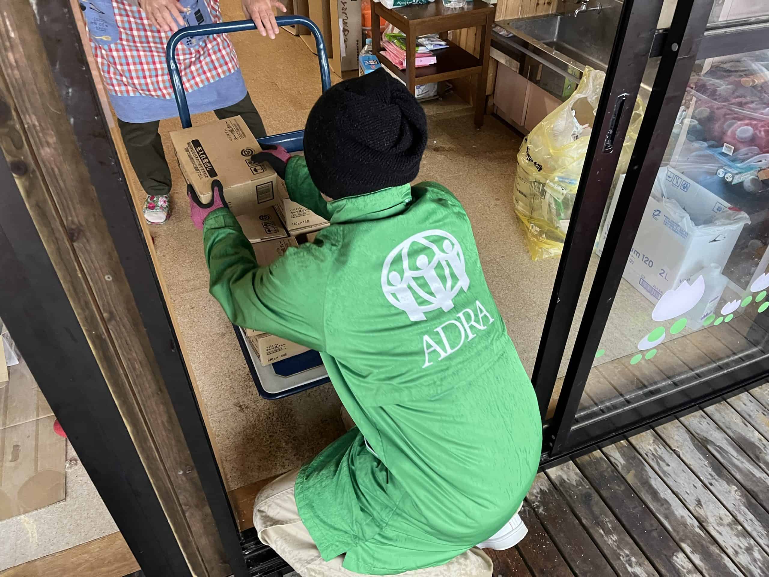 A person in a green jacket with the ADRA logo kneels down beside boxes