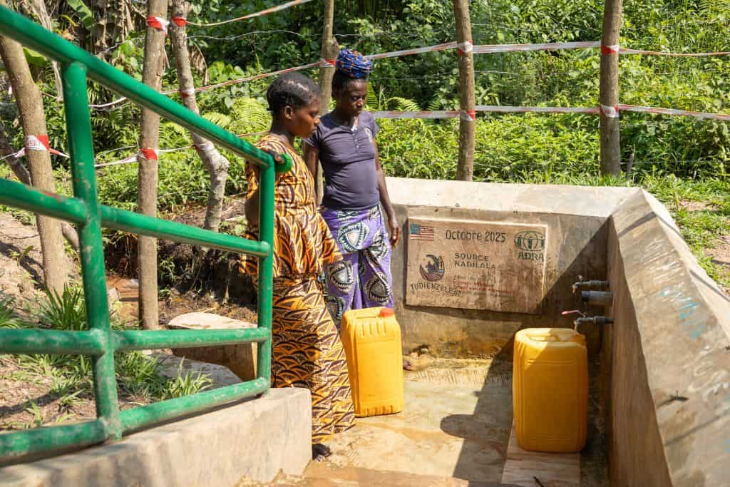 Two women from the Democratic Republic of the Congo stand are standing on a concrete platform waiting to fill their water buckets up.