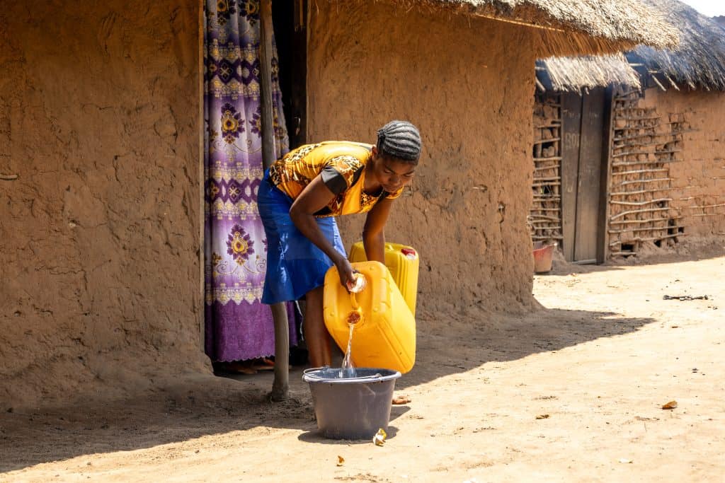 A girl from the Democratic Republic of the Congo is wearing a bright yellow top. She is pouring water into a bucket in front of her house.
