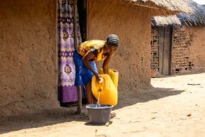 A girl from the Democratic Republic of the Congo is wearing a bright yellow top. She is pouring water into a bucket in front of her house.