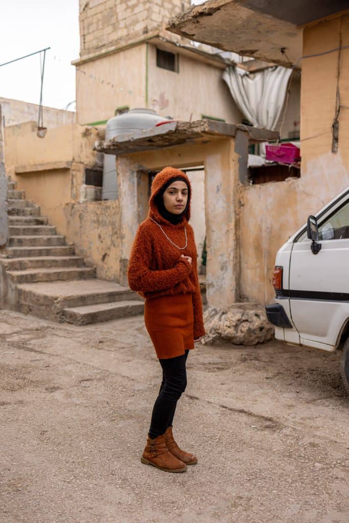 A woman standing in a poverty-stricken neighborhood with makeshift housing and a car in the backgrou.