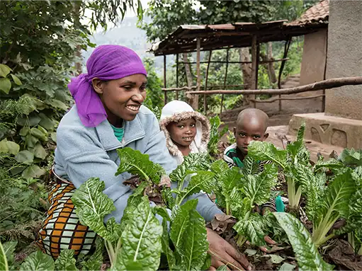 Woman and children gardening in a rural setting with lush green plants.