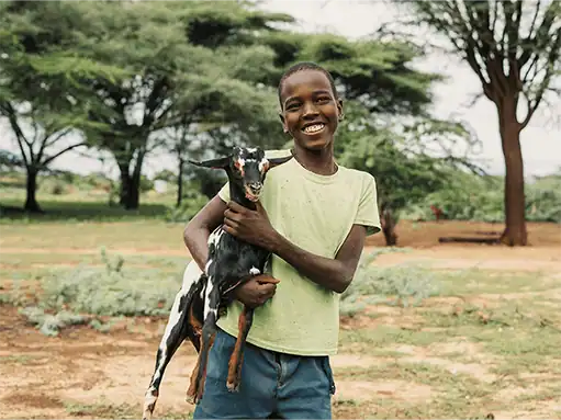 ADRA International, child holding goat in rural outdoor setting, smiling.