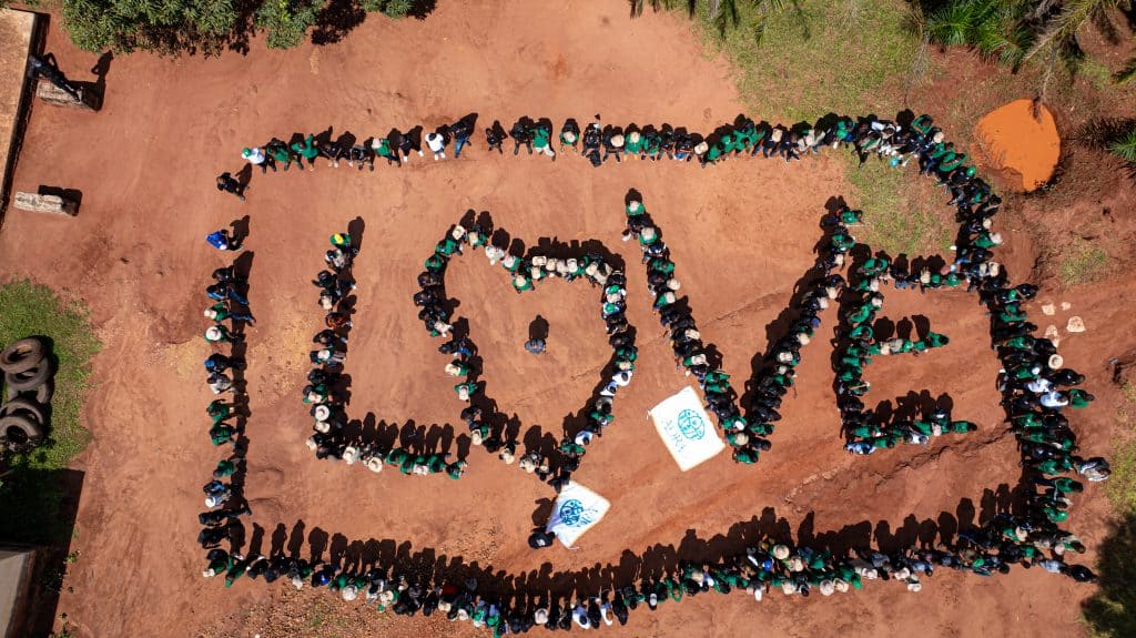 People forming the word "LOVE" with a heart symbol inside, outdoors on a dirt surface, during a comm.
