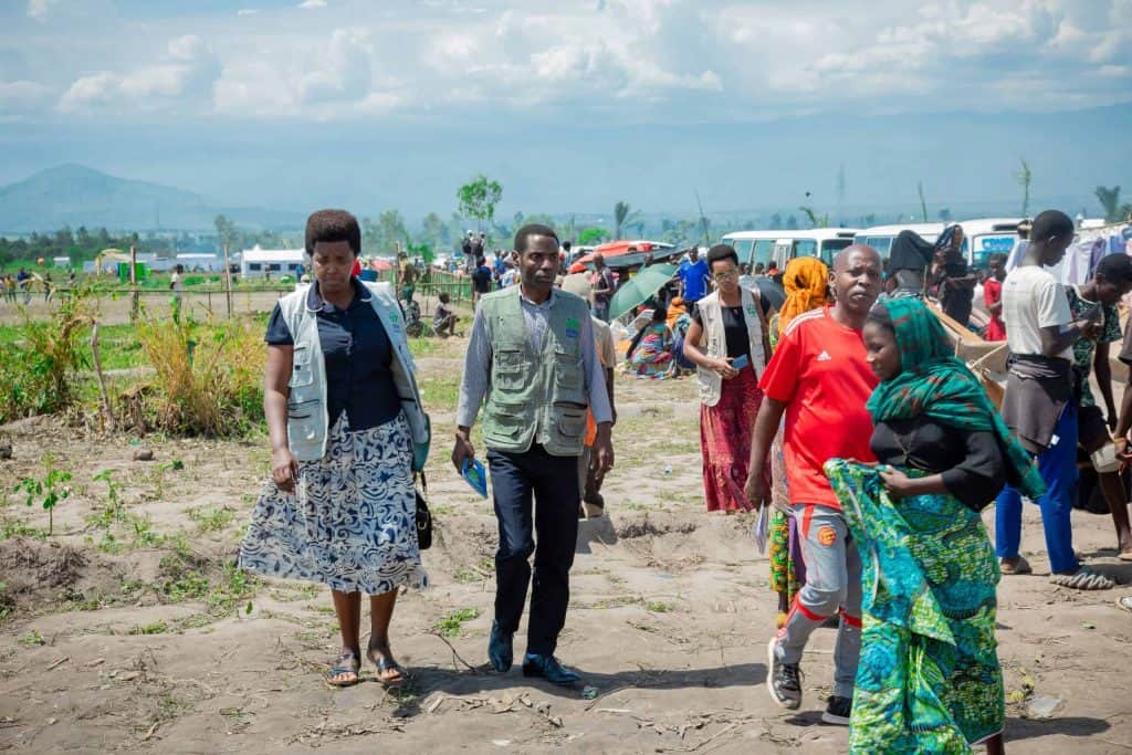 Refugees and aid workers in a refugee camp with tents and vehicles in the background.