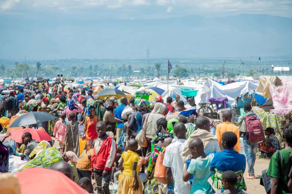 Large crowd of refugees with tents and umbrellas in a refugee camp.