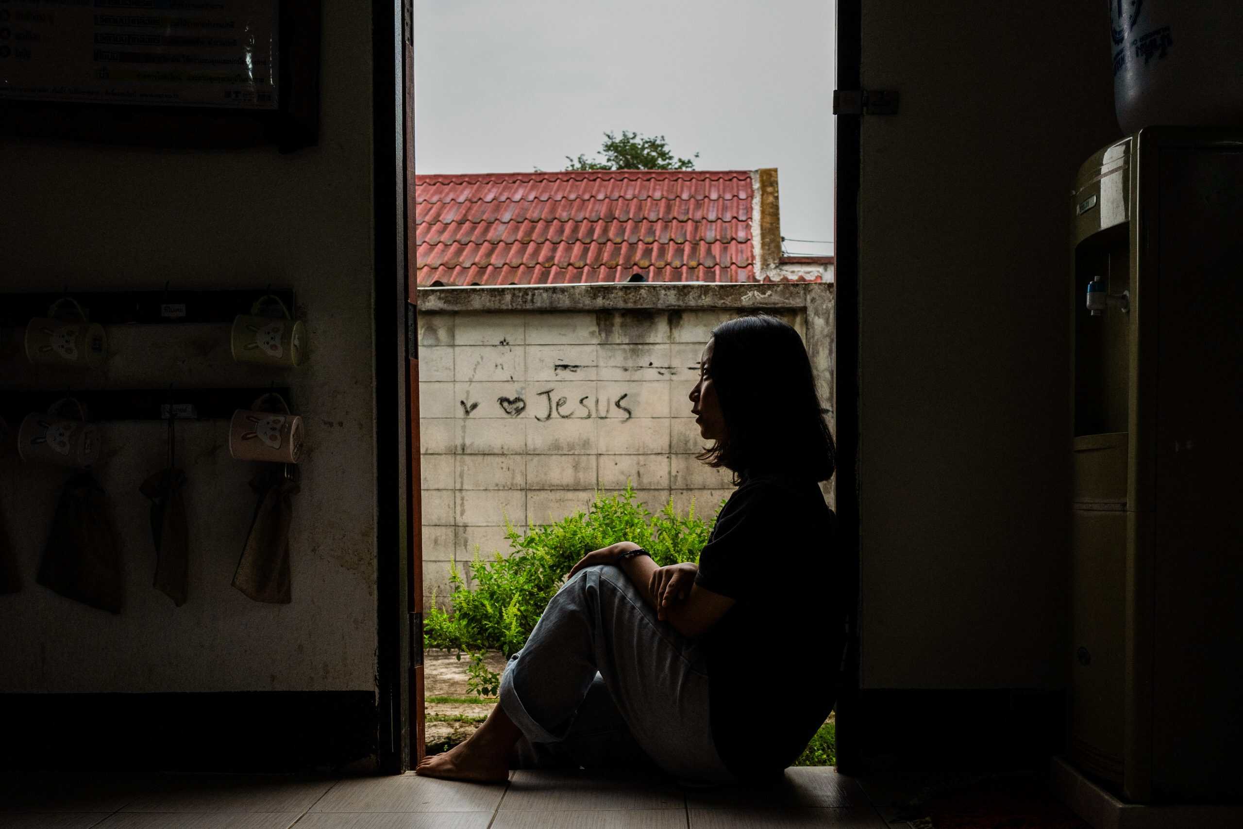 Silhouette of a woman sitting by an open door with 'I love Jesus' graffiti outside.
