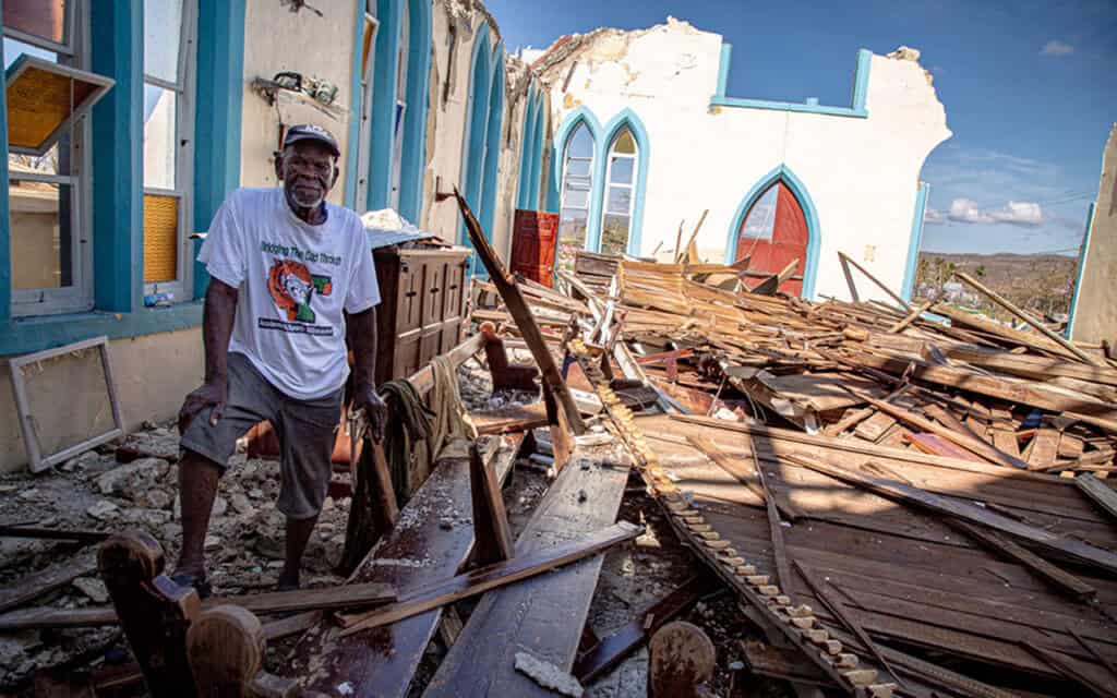 A man standing amidst the debris of a damaged building after a disaster, with partially destroyed wa.