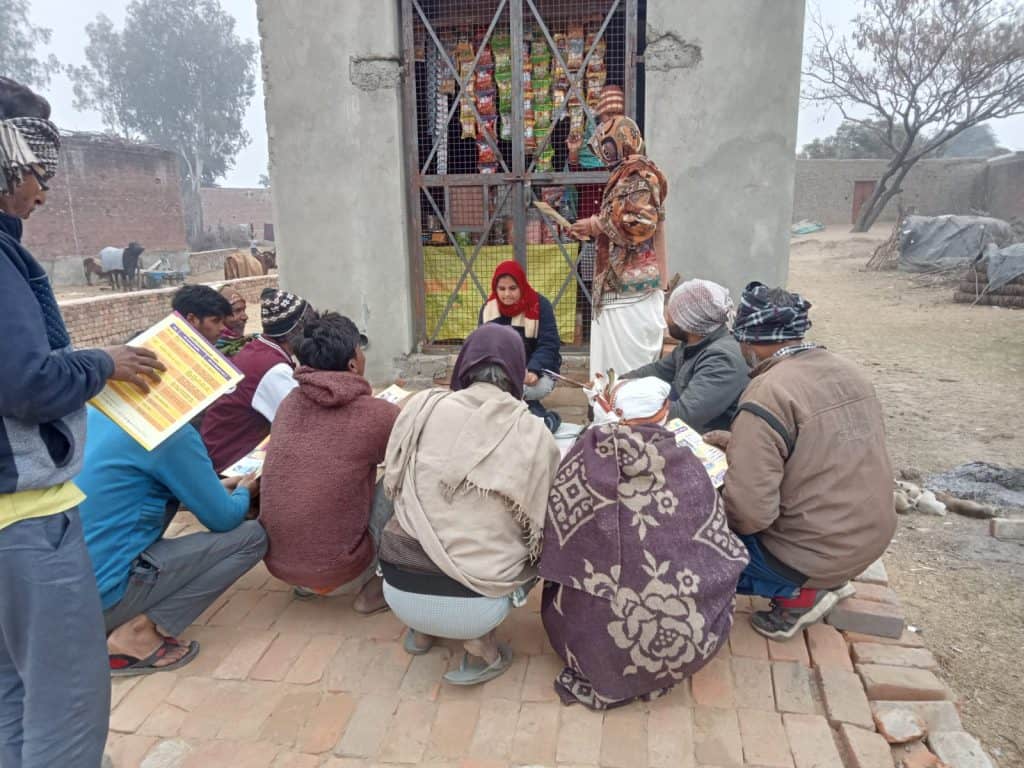 Community gathering with a woman speaking to seated villagers outside a building.