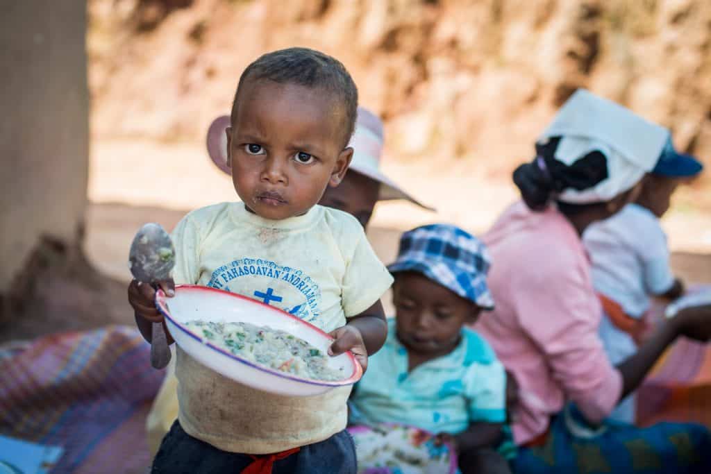 Young child holding a bowl of nutritious food in a community setting.