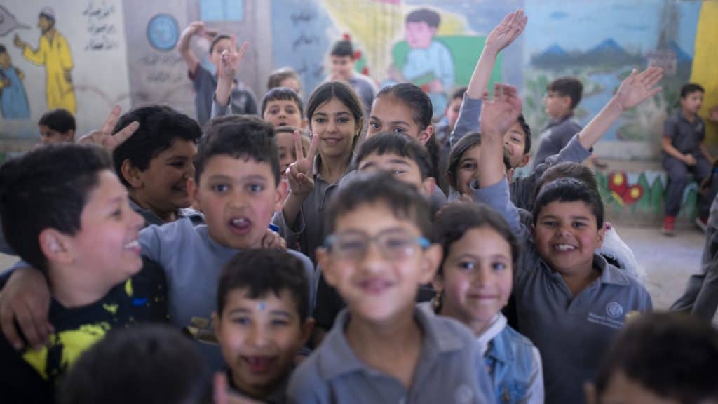 A group of smiling children in a classroom or community setting, engaging with the camera.