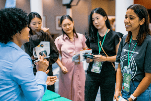 A young woman with curly hair and a blue shirt is standing a booth, talking to 4 other young women about ADRA